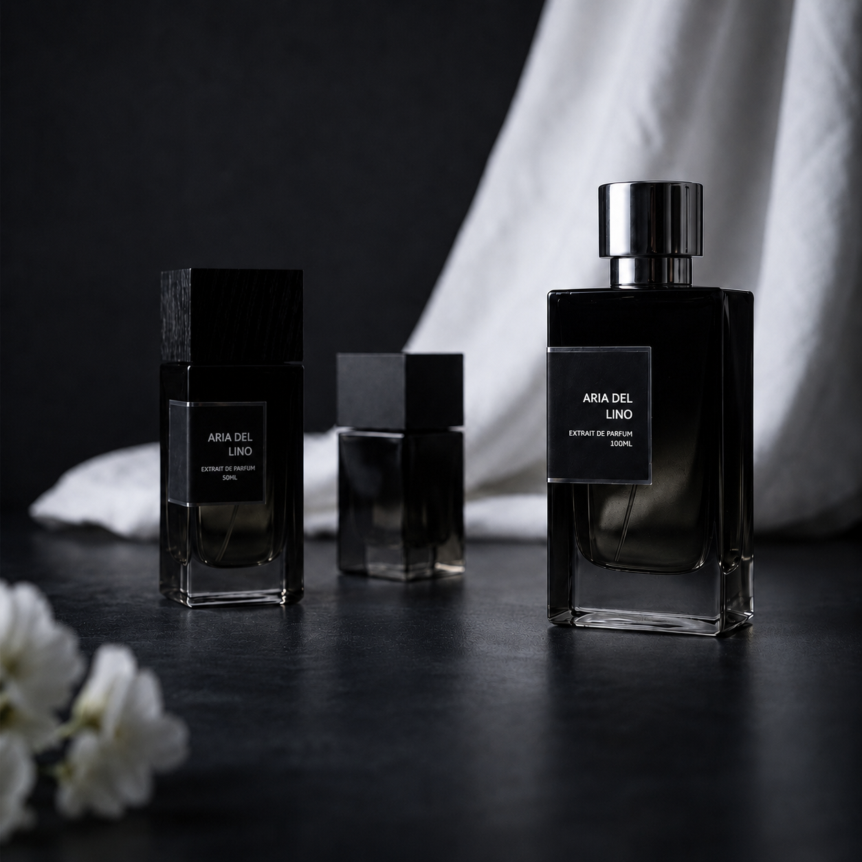 Three Aria del Lino perfume bottles on a dark surface with a white fabric backdrop and soft flowers in the foreground.